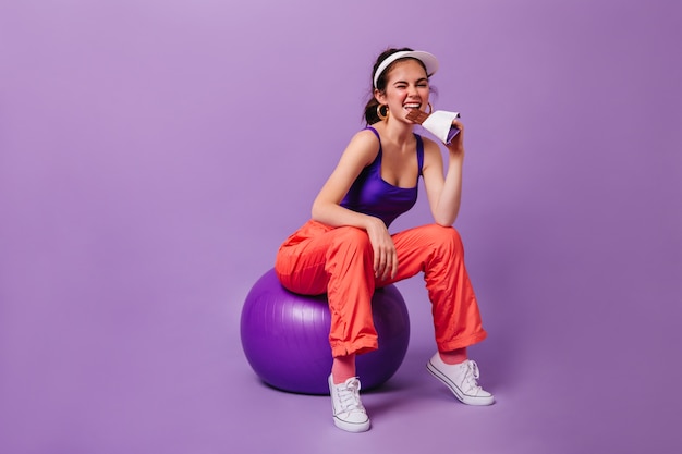 Mulher estilosa com blusa roxa e calça de moletom vermelha comendo barra de chocolate sentada em fitball contra a parede roxa