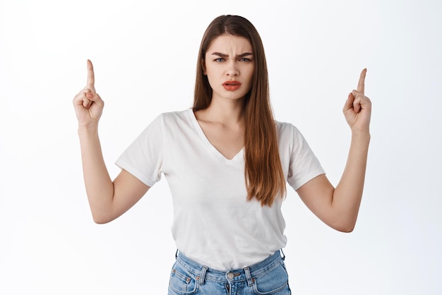 Mulher estilosa confusa franzindo a testa apontando para algo estranho olhar descontente e intrigado questionado pelo acordo promocional de pé na camiseta contra o fundo branco