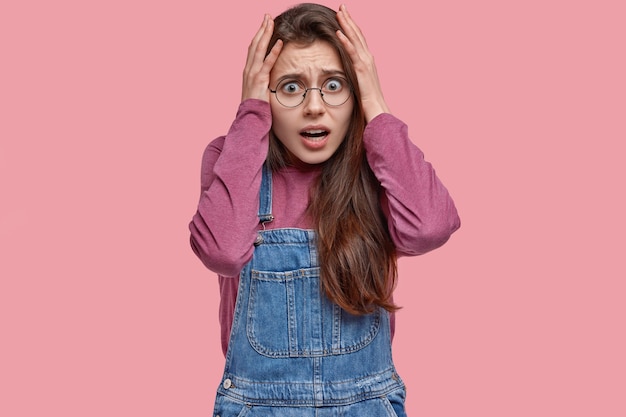 Mulher estressada fica com as mãos na cabeça em pânico, tem alguns problemas, olha com expressão assustada, vestida com roupas da moda