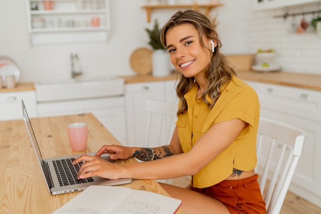 Mulher estudante bonita usando laptop e escrevendo notas em sua cozinha moderna e leve.