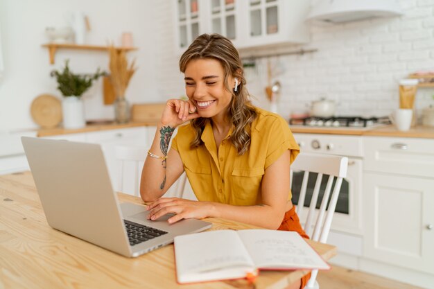 Mulher estudante bonita usando laptop e escrevendo notas em sua cozinha moderna e leve.