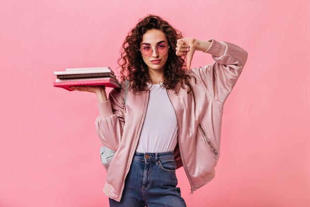 Mulher estudante usando óculos de sol rosa segurando livros e mostrando o polegar para baixo
