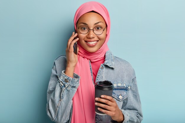 Mulher étnica alegre de aparência agradável tem uma conversa casual agradável, desfruta da pausa matinal para o café, segura um copo descartável de bebida, usa óculos redondos, cobre a cabeça com hijab. Conceito de tempo livre