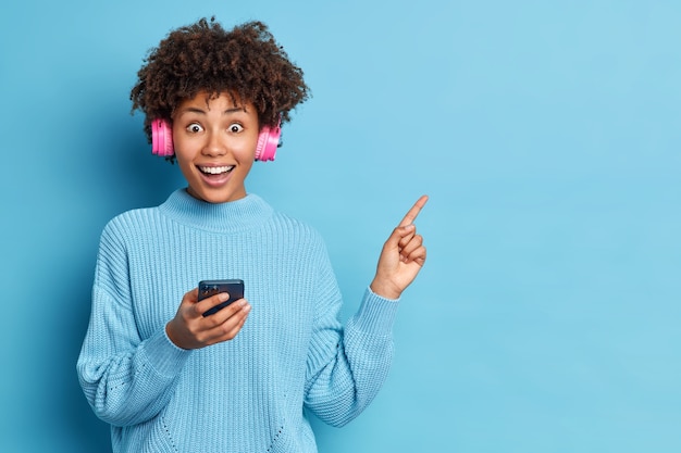 Mulher étnica feliz com cabelo afro indica no canto superior direito segurando um celular moderno e ouvindo música por meio de fones de ouvido estéreo vestindo um suéter de tricô solto