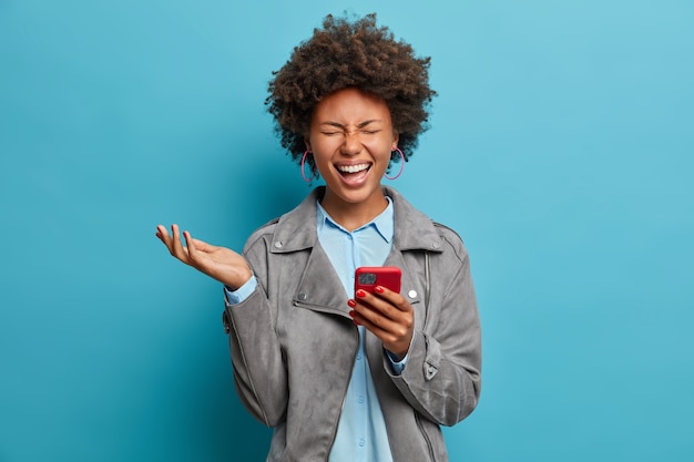 Mulher étnica radiante com cabelos cacheados, ri enquanto assiste a um vídeo engraçado no smartphone, fecha os olhos e levanta a mão, estando de bom humor, usa uma jaqueta casual cinza, poses