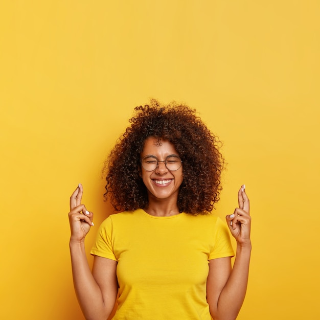 Mulher étnica supersticiosa alegre espera pelo melhor, cruza o dedo para dar sorte, ansiosa para conseguir uma posição em uma grande empresa, sorri com alegria, usa óculos e camiseta, isolada na parede amarela.