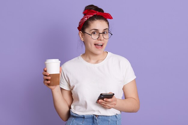 Mulher europeia alegre alegre em uma camiseta casual branca e uma faixa vermelha, segurando um café para viagem e navegando na internet no celular, mandando mensagem para um amigo,