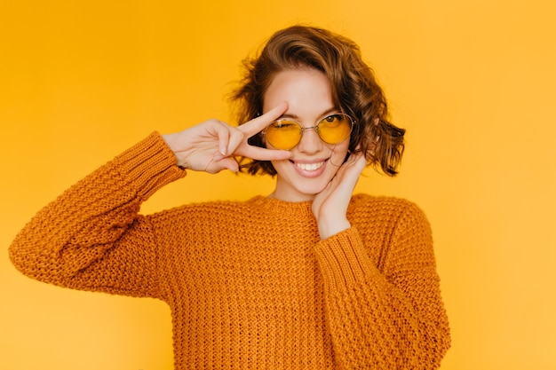 Mulher europeia alegre com cachos brilhantes rindo e mostrando o símbolo da paz em fundo amarelo