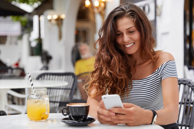 Mulher européia atraente com expressão alegre usa celular moderno em uma cafeteria aconchegante