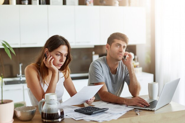 Mulher européia atrativa nova que senta-se na mesa de cozinha ao lado de seu marido, lendo atentamente o contrato, calculando figuras com calculadora. Homem de negócios fazendo bonito chamada por telefone celular