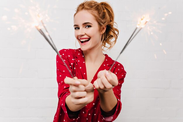 Mulher europeia bonita que comemora as férias de inverno com luzes de Bengala. Agradável menina caucasiana de pijama vermelho segurando estrelinhas e sorrindo.