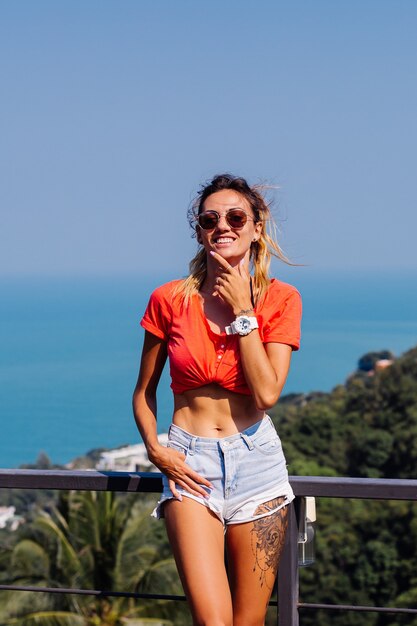 Mulher européia caucasiana tatuada e bronzeada com corpo e abdômen esportivos, shorts jeans e camisa laranja vermelha