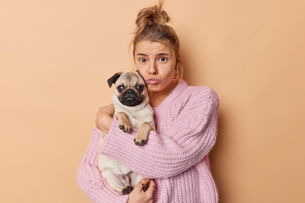 Mulher europeia chateada olha tristemente para a câmera abraça o cão pug com amor se sente infeliz por causa de problemas bolsas lábios usa suéter de malha isolado sobre fundo marrom Conceito de pessoas e animais de estimação