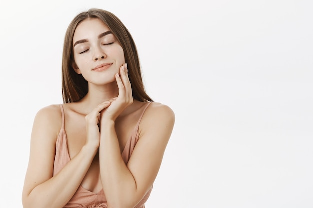 Mulher européia de boa aparência gentil e terna com lindas características faciais fechando os olhos e sorrindo sensualmente, tocando a pele delicadamente e se deliciando com o produto para a pele