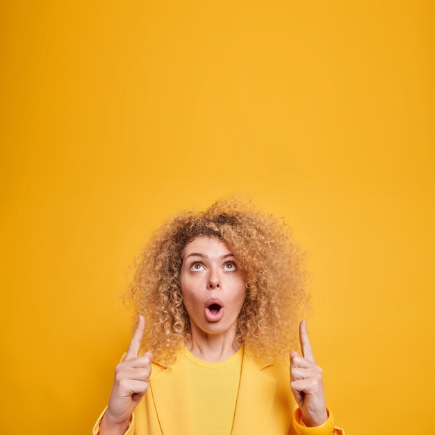 Mulher européia de cabelos cacheados surpresa e fascinada pontos acima impressionada com promo incrível mantém a boca aberta vestida elegantemente isolada sobre parede amarela mostra interessante negócio de compras.