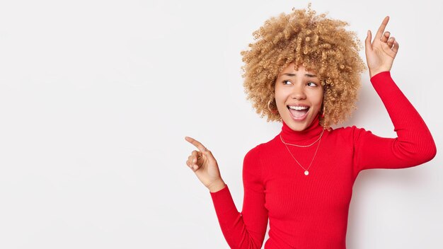 Mulher europeia de cabelos encaracolados alegre enérgica vestida de gola alta vermelha dança despreocupada canta música tem poses de humor otimista contra fundo branco espaço em branco à esquerda para o seu conteúdo promocional