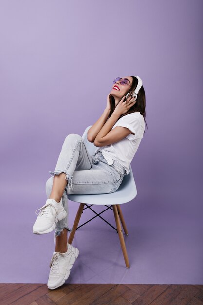 Mulher europeia encantadora de tênis branco, ouvindo música. Retrato de uma garota feliz com cabelo castanho, sentado na cadeira com fones de ouvido.