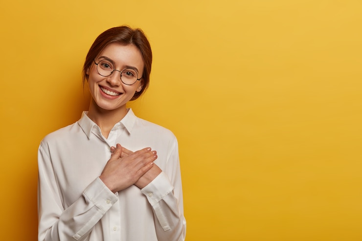 Foto grátis mulher europeia encantadora e positiva aperta as mãos contra o peito, expressa gratidão pelo presente, agradece a ajuda, usa óculos redondos e camisa branca