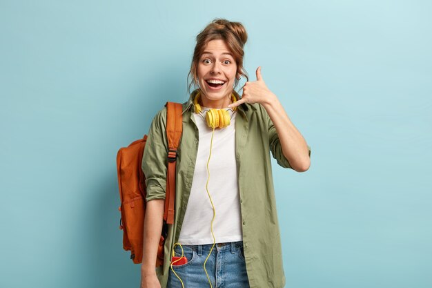 Mulher europeia feliz faz gesto de chamada, tenta fazer contato com um amigo à distância, usa fones de ouvido modernos