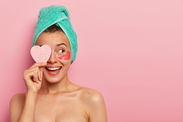 mulher européia feliz tem sorriso largo, enxuga os olhos com esponja em formato de coração, tira maquiagem, tem corpo e pele bem cuidados