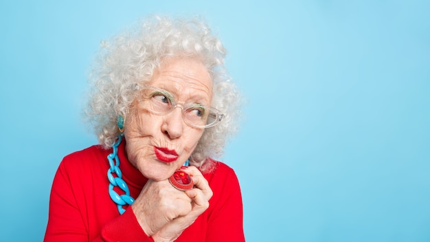 Mulher europeia idosa, sonhadora e pensativa, mantém os lábios arredondados de lado e pensa em algo agradável vestida com roupas da moda