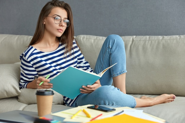 Mulher europeia pensativa de óculos transparentes, segura um livro e uma caneta, escreve anotações como se prepara para passar o trabalho do curso, bebe café para viagem, posa no sofá do apartamento. conceito de trabalho