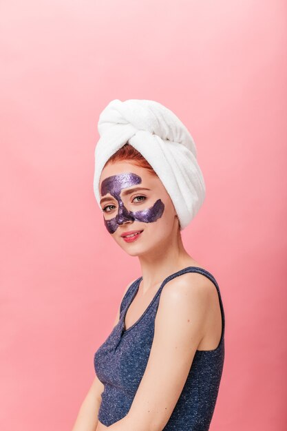 Mulher europeia refinada posando com máscara facial. Foto de estúdio de menina caucasiana com uma toalha na cabeça.