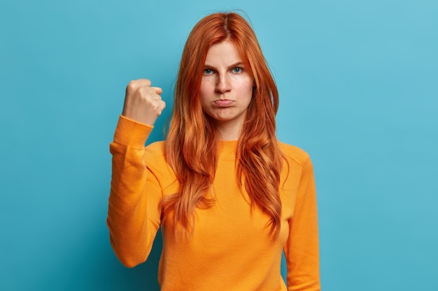 Mulher europeia ruiva séria parece com raiva mostra punho pede para não incomodar os lábios da bolsa e tem expressão facial irritada vestida com blusão laranja casual.
