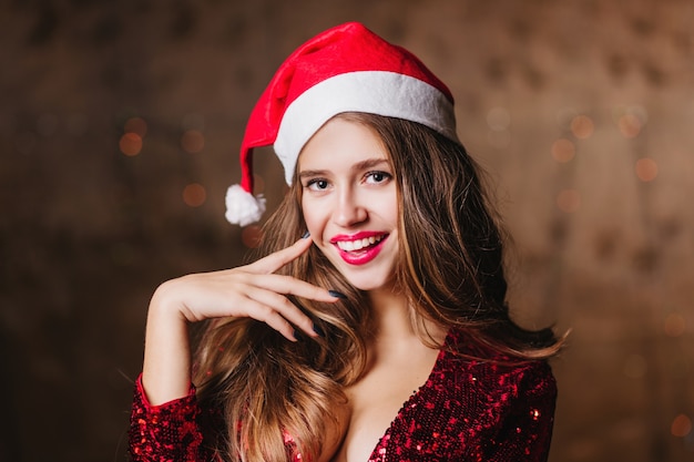 Mulher europeia sensual com chapéu de Papai Noel posando na parede escura e sorrindo