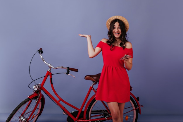 Mulher excitada com o telefone na mão, em pé ao lado de sua bicicleta. Menina morena emocional com chapéu de palha, posando na frente da bicicleta.