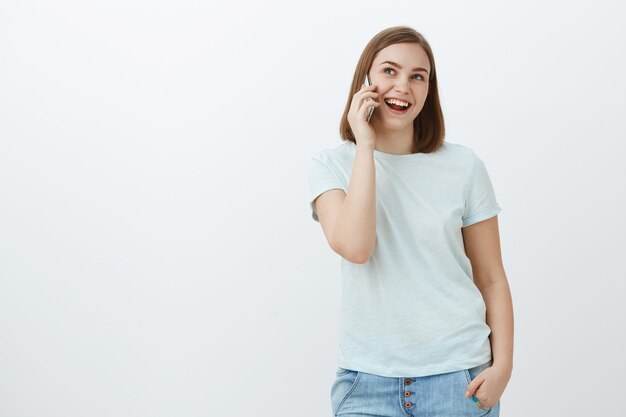 Mulher falando alegremente com um amigo pode ficar horas no telefone. Mulher sociável, feliz e descontraída, bonita, de camiseta, jeans segurando a mão no bolso e espertinho perto da orelha olhando para o lado com um sorriso