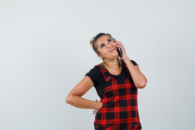 Mulher falando no celular com vestido de avental e olhando pensativa, vista frontal.