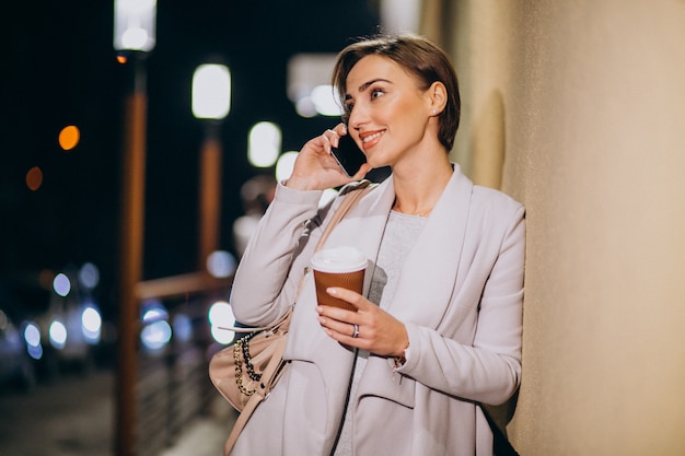 Mulher falando no telefone e tomando café lá fora na rua à noite