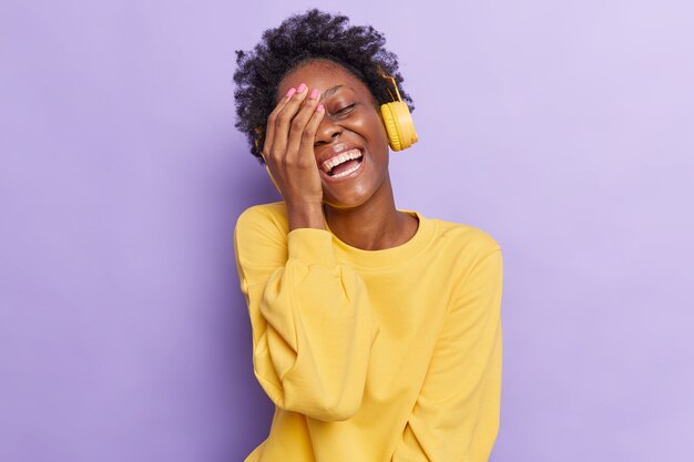 mulher faz o rosto com a palma da mão sorri amplamente de algo muito engraçado ouve música pelos fones de ouvido vestida com um macacão amarelo isolado no roxo