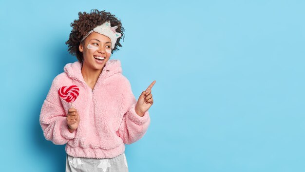 Mulher feliz apontando para o espaço da cópia sente-se energizada pela manhã, depois que um bom sono indica de lado e anuncia produto para uma ótima soneca segura pirulito usa pijama isolado sobre parede azul