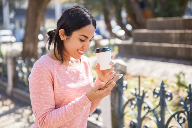 Mulher feliz bebendo café e navegando no smartphone ao ar livre