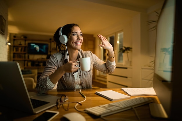 Mulher feliz bebendo chá e acenando para alguém enquanto faz videochamada no PC de mesa à noite em casa