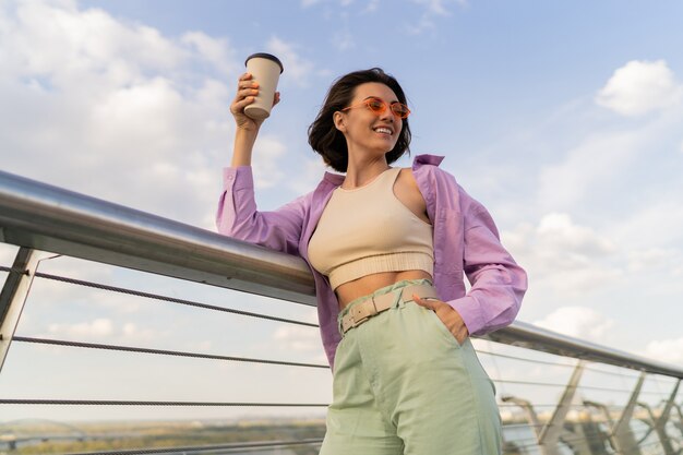 Mulher feliz com a figura perfeita em uma elegante camisa roxa grande, desfrutando de uma xícara de café enquanto caminha na ponte moderna