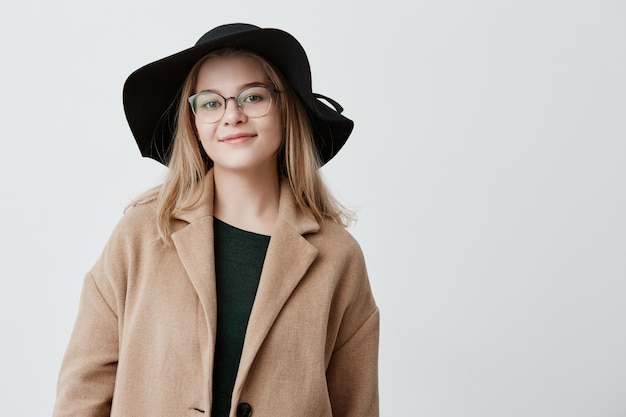 Mulher feliz com aparência atraente, vestida com casaco da moda e chapéu preto, com óculos, expressa emoções positivas, descansa depois de passear no parque com os amigos, tem bom humor.