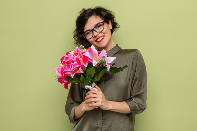 Mulher feliz com cabelo curto segurando um buquê de flores olhando para a câmera sorrindo alegremente, comemorando o dia internacional da mulher, 8 de março, em pé sobre fundo verde