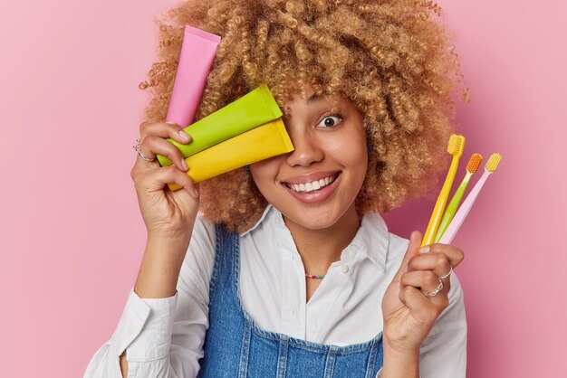 Mulher feliz com cabelo encaracolado segura escovas de dentes e tubos de creme dental sorri com prazer tem dentes bem cuidados usa camisa branca e jeans sarafan isolado sobre fundo rosa Conceito de atendimento odontológico