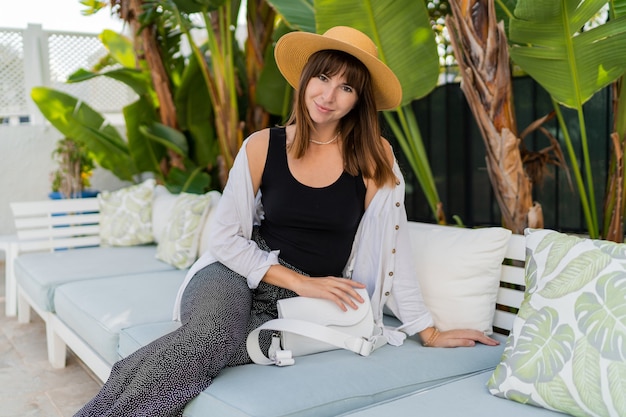 Mulher feliz com chapéu de palha relaxando em casa, no terraço de luxo, posando perto de jardim tropical.