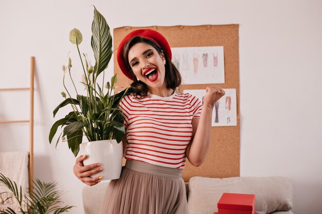 Mulher feliz com lábios vermelhos, segurando a planta. Menina na moda com chapéu brilhante, camiseta listrada e saia bege se alegra no apartamento.
