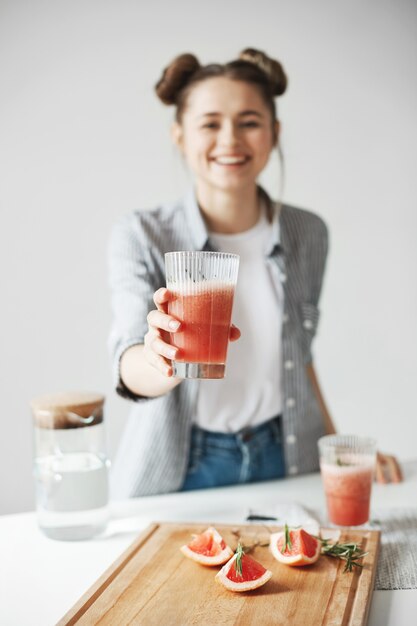 Mulher feliz com os bolos que sorriem esticando o batido da desintoxicação da toranja sobre a parede branca. Comida de dieta saudável. Concentre-se no vidro.