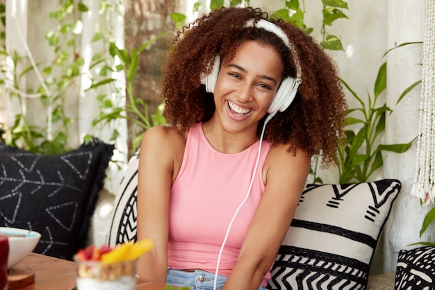 Mulher feliz com pele escura e saudável, ouve anedotas online com aplicativos especiais e fones de ouvido, ri de uma piada engraçada, senta-se no sofá confortável contra o aconchegante interior do café. Pessoas e tempo livre