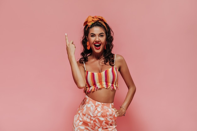 Mulher feliz com penteado preto encaracolado e bandana laranja em um top moderno e calças rosa legais aparecendo e olhando para a câmera.