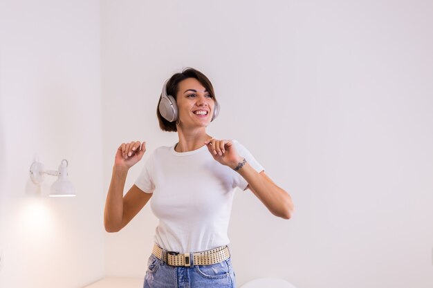 Mulher feliz com roupas casuais em casa no quarto ouvindo música em fones de ouvido, dançando e pulando