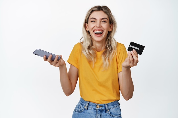 Mulher feliz com smartphone e cartão de crédito rindo pagando por algo ganhando dinheiro no telefone em pé contra fundo branco