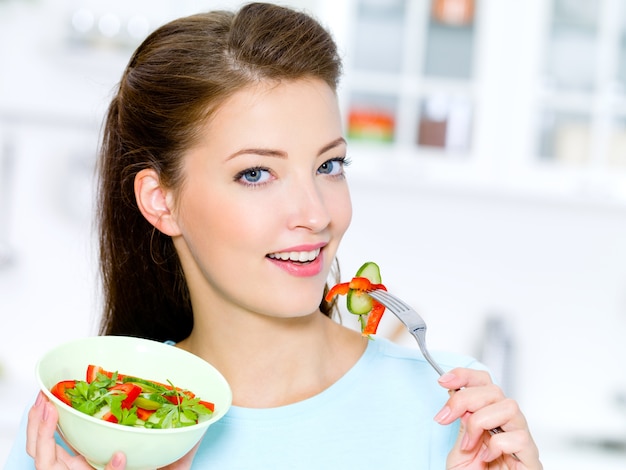 Mulher feliz comendo salada de legumes na cozinha