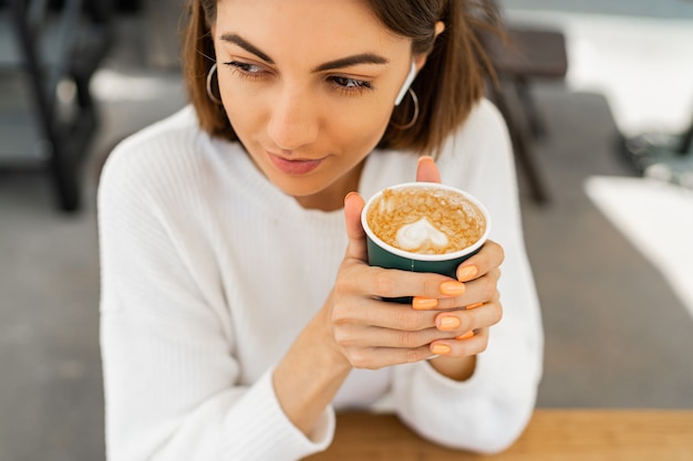 Mulher feliz de cabelos curtos saboreando capuccino em um café, vestindo um suéter branco aconchegante